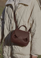 Person wearing a beige quilted jacket with a brown leather backpack in an outdoor setting