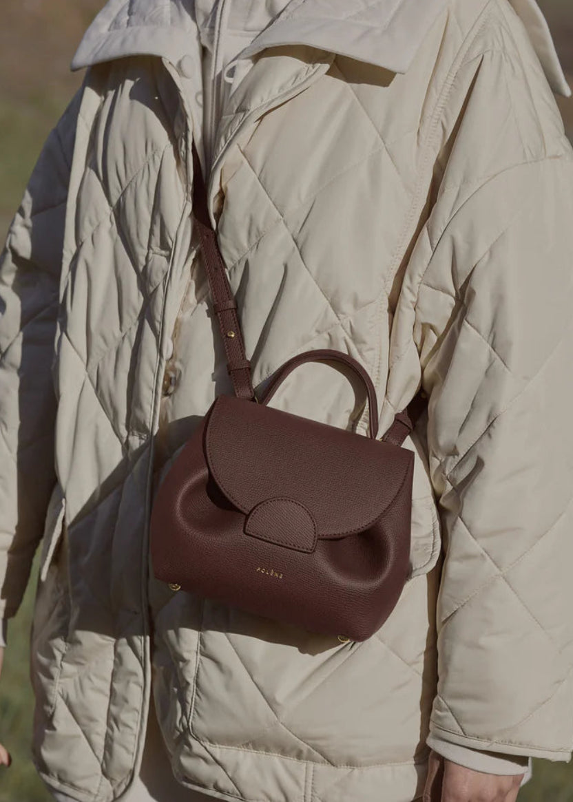Person wearing a beige quilted jacket with a brown leather backpack in an outdoor setting